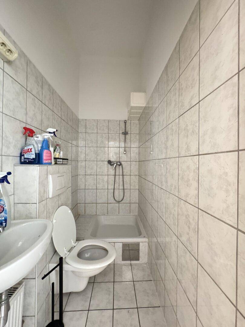 Kleines Badezimmer mit weißen Fliesen, Toilette, Waschbecken und Duschbereich.