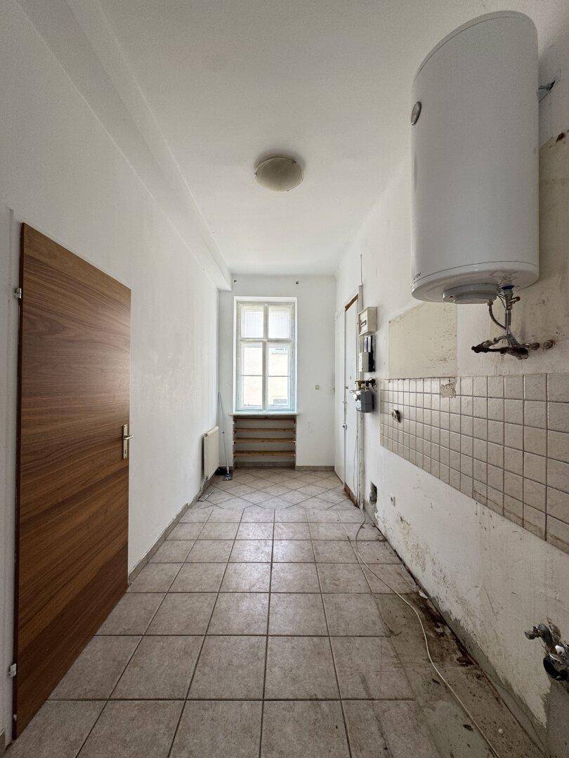 Langer Flur mit Fliesenboden, Fenster am Ende und Warmwasserboiler an der Wand.