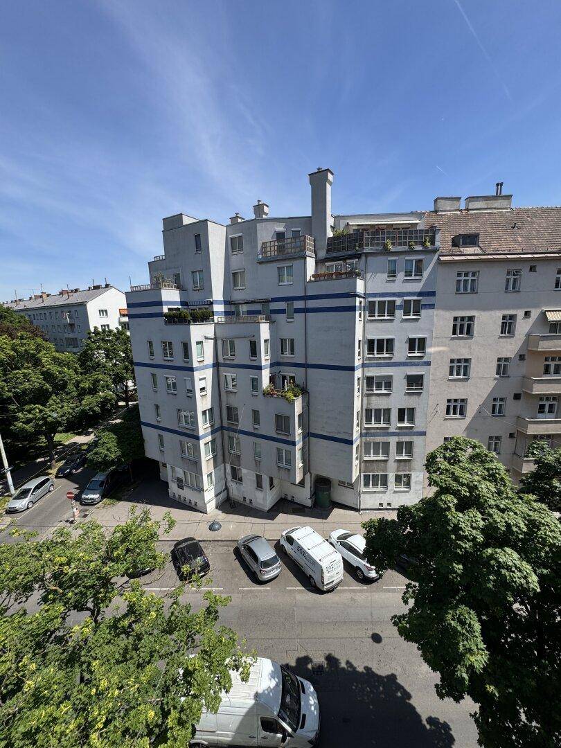 Panoramablick auf eine belebte Stadtstraße mit parkenden Autos, Bäumen und Wohngebäuden.