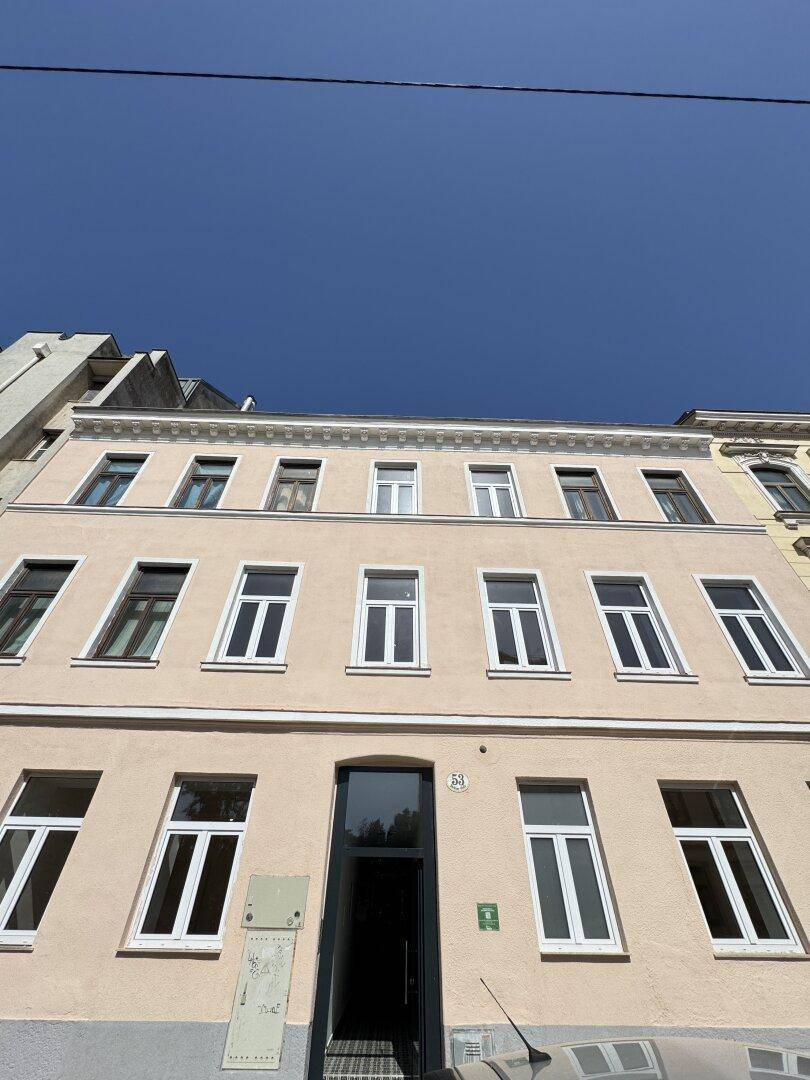 Fassade eines mehrstöckigen Gebäudes mit vielen Fenstern und einem Eingang unter blauem Himmel.