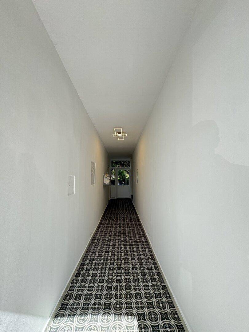 Langer, schmaler Flur mit gemustertem Fliesenboden und moderner Deckenleuchte, der zum Ausgang führt.