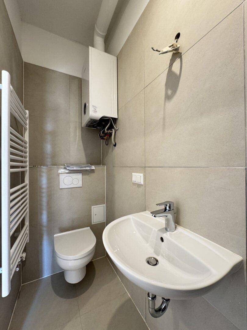 Kleines WC mit Wand-WC, Waschbecken, Handtuchheizkörper und einem Warmwasserbereiter.