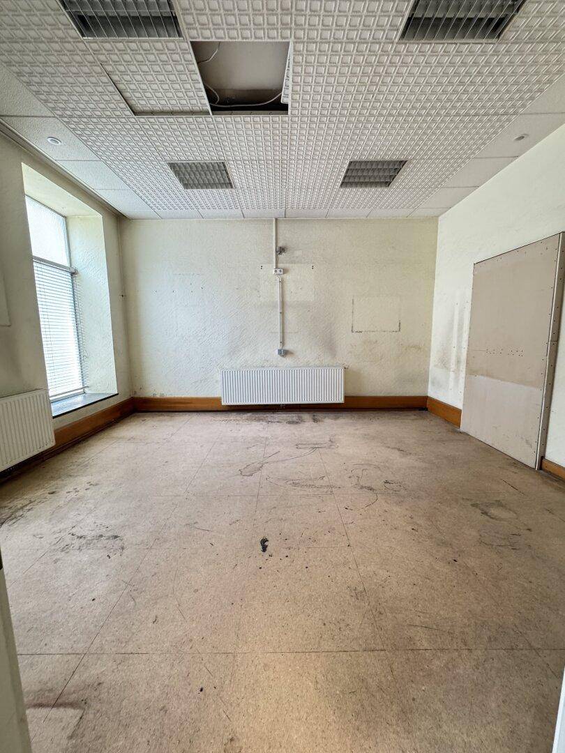 Leerer Raum mit abgehängter Decke, großem Fenster und Spuren von Abnutzung auf dem Boden.