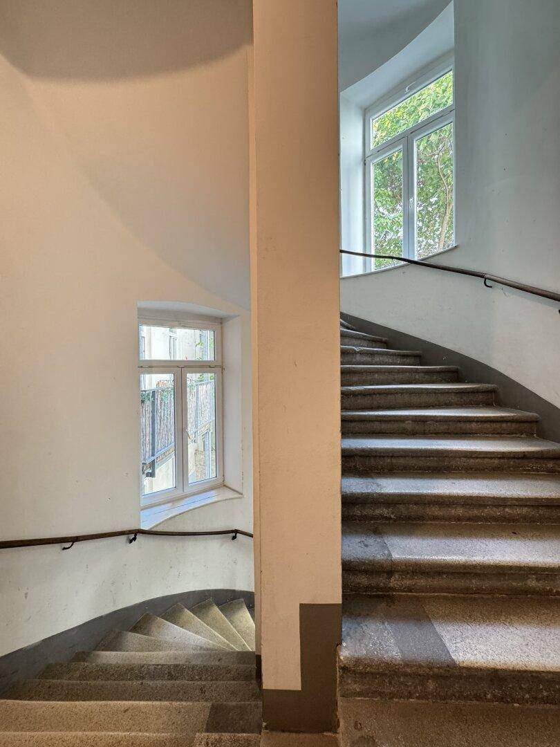 Historisches Treppenhaus mit Steinstufen und großen Fenstern, die viel Licht hereinlassen.
