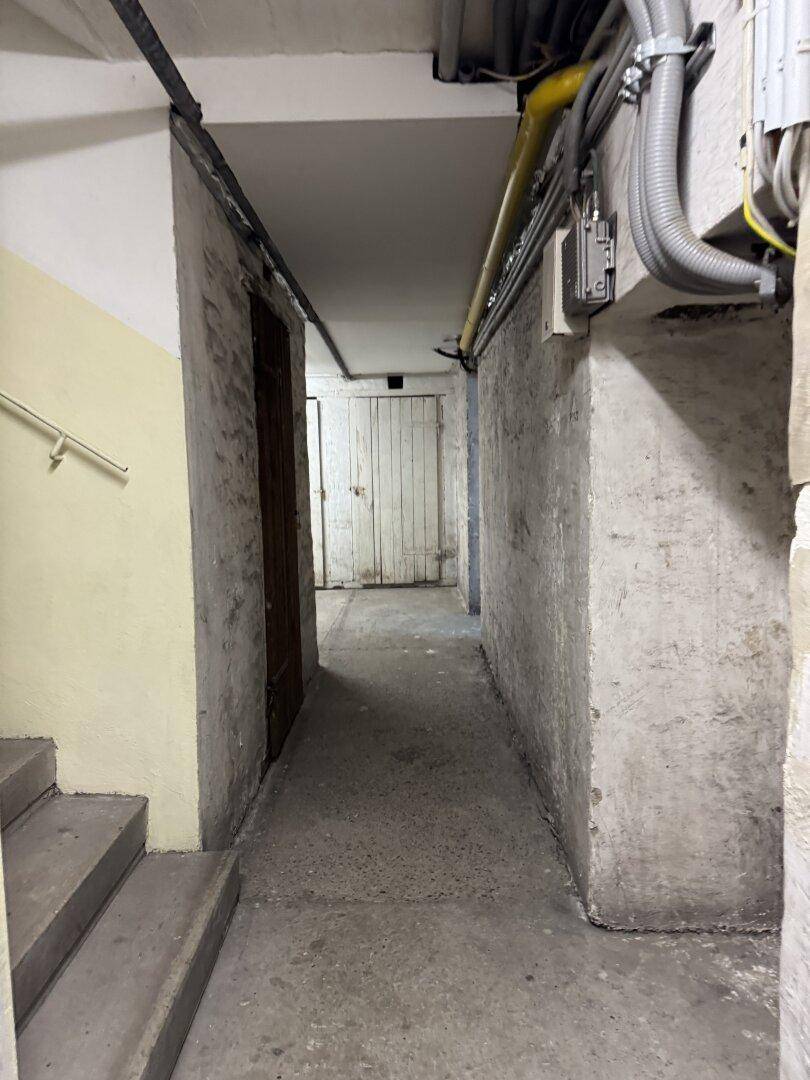 Langer Kellergang mit Lagermöglichkeiten und sichtbaren Versorgungsleitungen an der Decke.