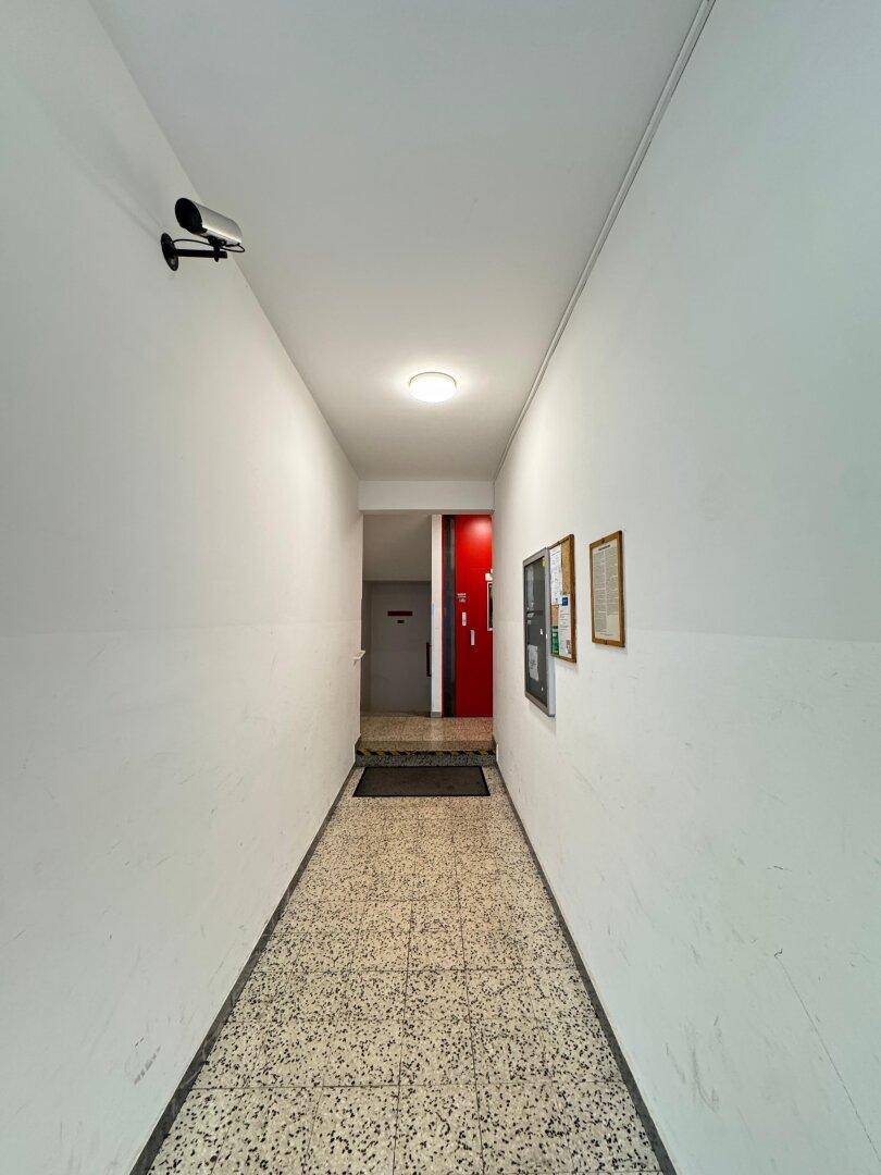 Langer, heller Flur im Gebäude mit Terrazzoboden und Überwachungskamera.
