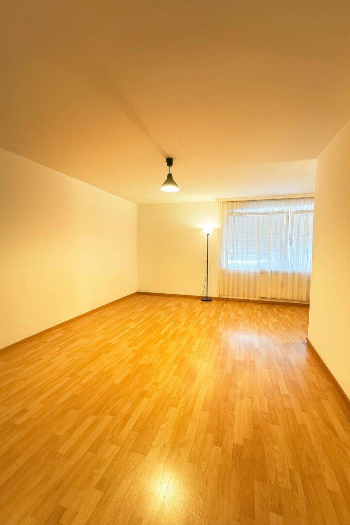 Heller, leerer Raum mit Parkettboden, großem Fenster und einer Stehlampe.