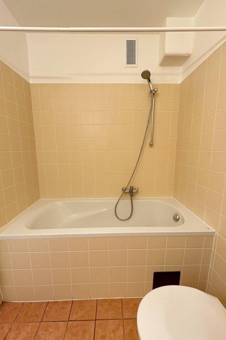 Badezimmer mit Badewanne, beigefarbenen Fliesen und Duschvorrichtung.