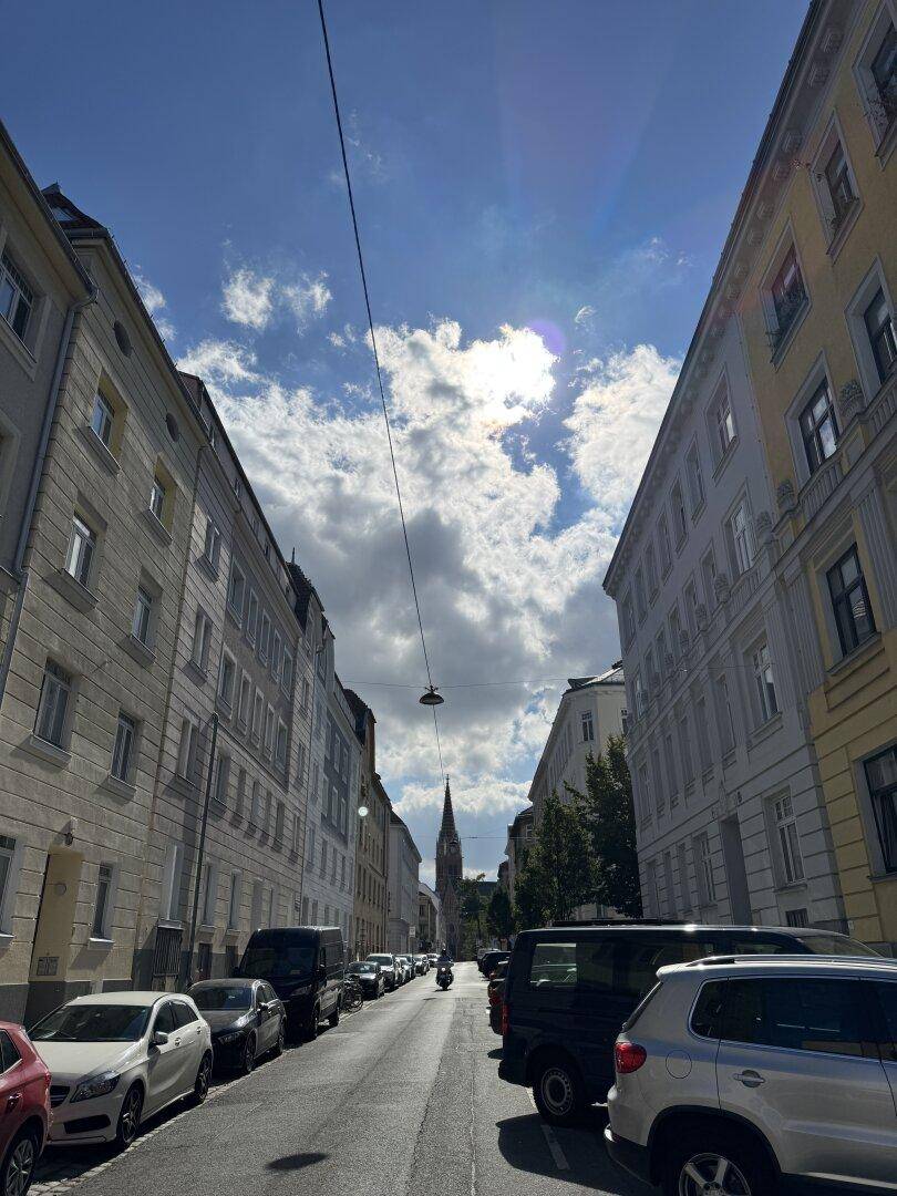 Stadtstraße mit parkenden Autos und Gebäuden, im Hintergrund ist ein Kirchturm zu sehen.