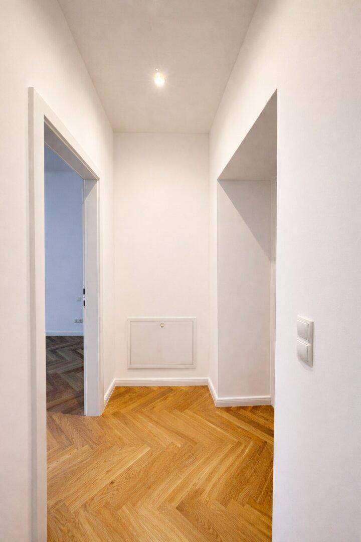 Heller Flur mit Fischgrätparkettboden und weiß gestrichenen Wänden, der zu weiteren Räumen führt.