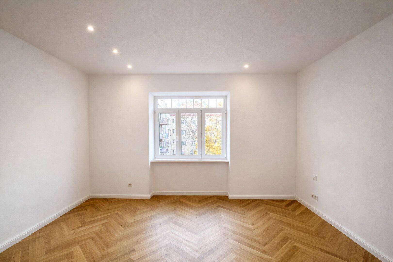 Lichtdurchflutetes Zimmer mit Fischgrätparkett und breitem Fenster, das viel Tageslicht hereinlässt.