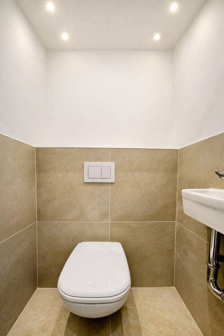 Separates WC mit wandhängender Toilette, beigefarbenen Fliesen und kleinem Waschbecken.