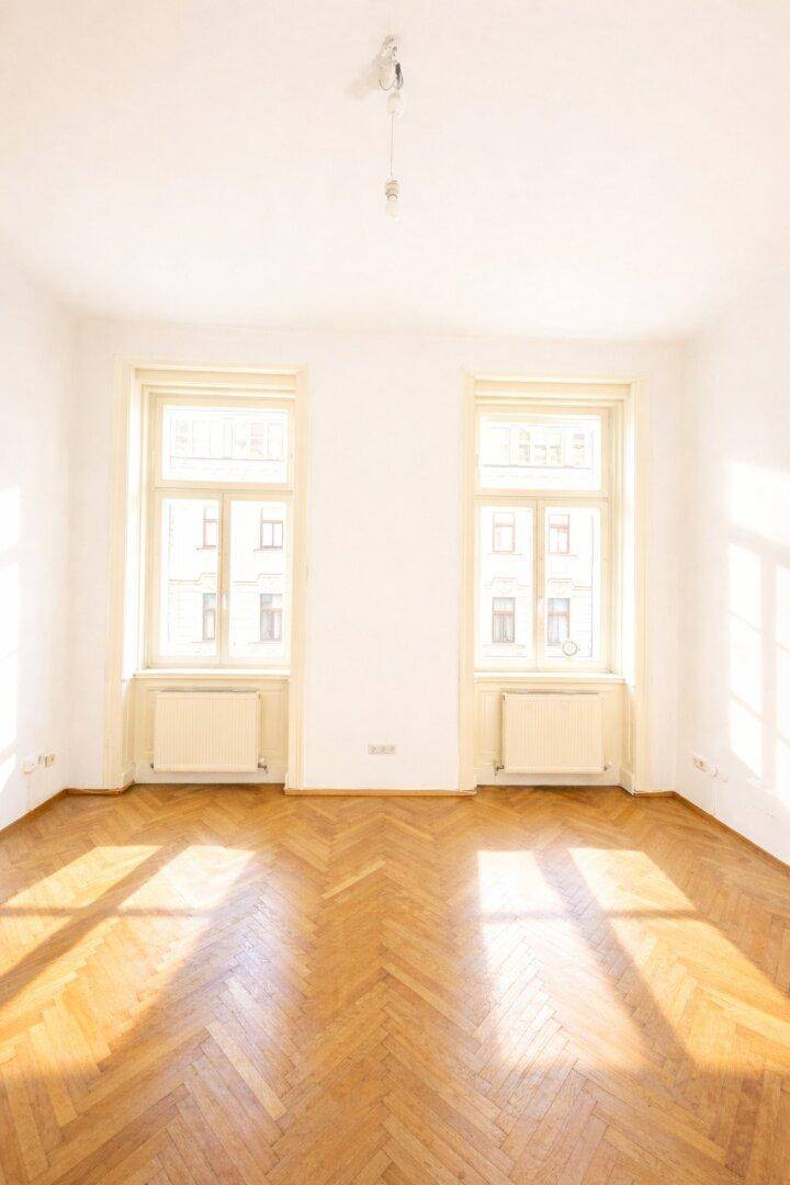 Lichtdurchfluteter Raum mit zwei großen Fenstern und elegantem Fischgrätparkettboden, der viel Sonnenlicht einfängt.
