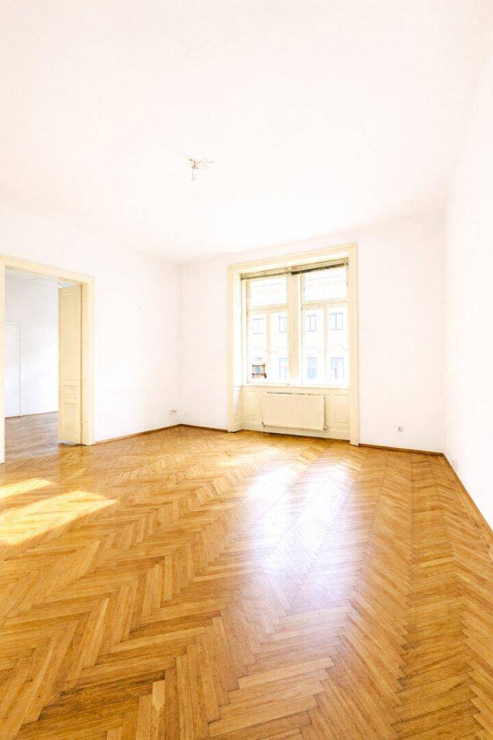 Geräumiges, leeres Zimmer mit klassischem Fischgrätparkett und einem Fenster mit Blick nach draußen.