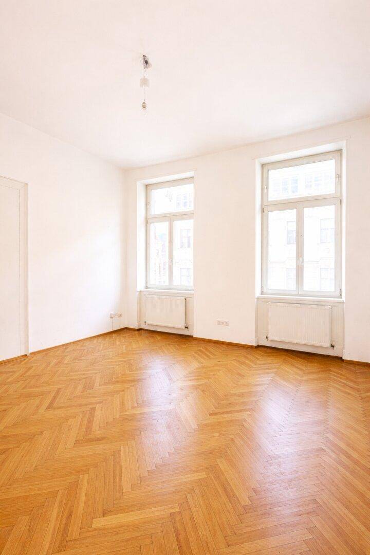 Leeres Zimmer mit hellem Fischgrätparkett und zwei Fenstern, die den Raum mit Tageslicht füllen.