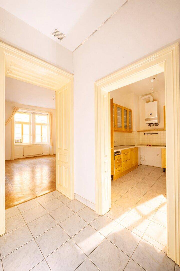 Flur mit hellen Fliesen, der zu einem Zimmer mit Parkett und einer Küche mit Einbauten führt.
