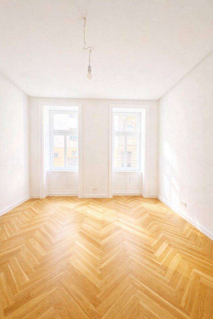 Helles, leeres Zimmer mit Fischgrätparkettboden und zwei großen Fenstern, bereit zur Einrichtung.