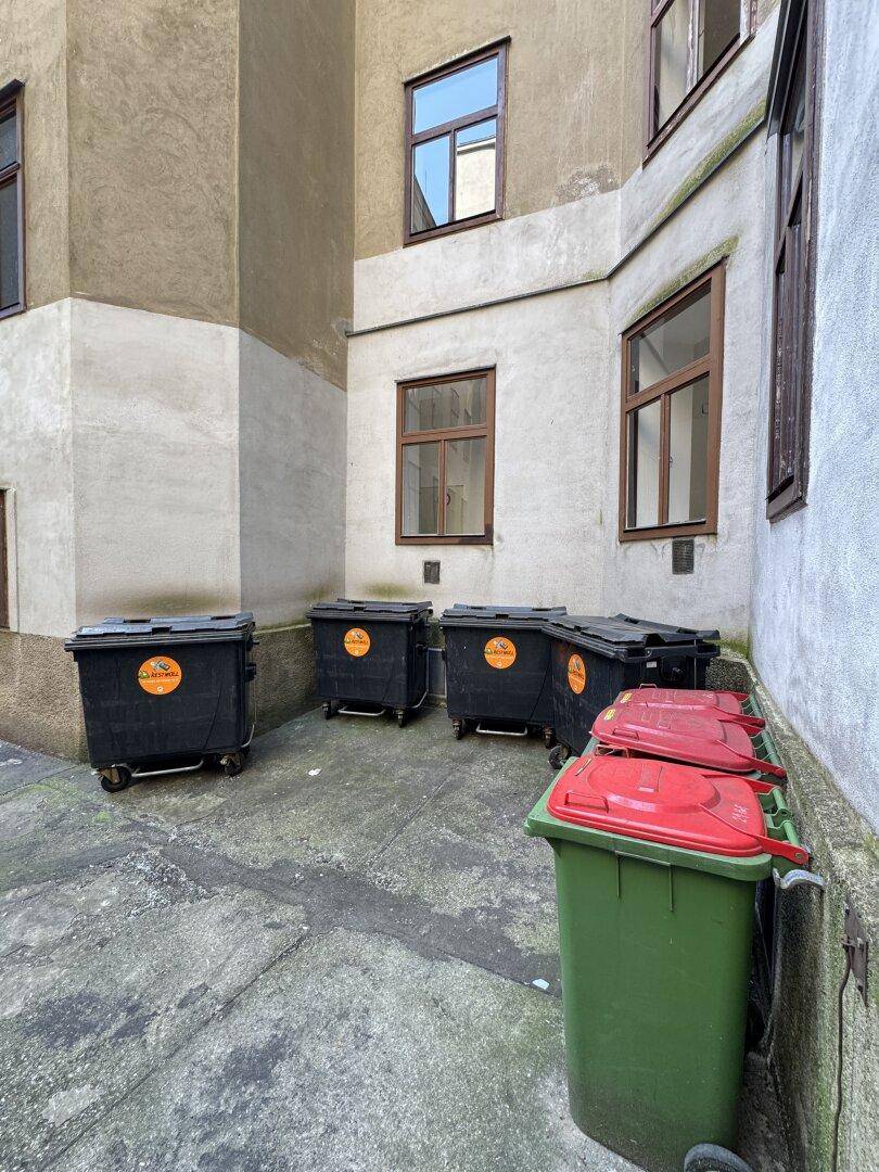 Gebäudeecke mit mehreren Mülltonnen im Außenbereich, Teil des Hinterhofs.