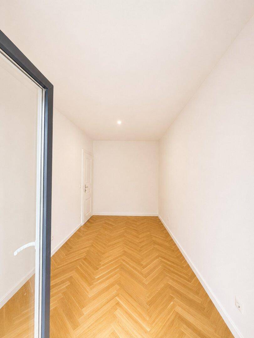 Helles Zimmer mit Fischgrätparkett und einer geöffneten Tür, die Licht hereinlässt.