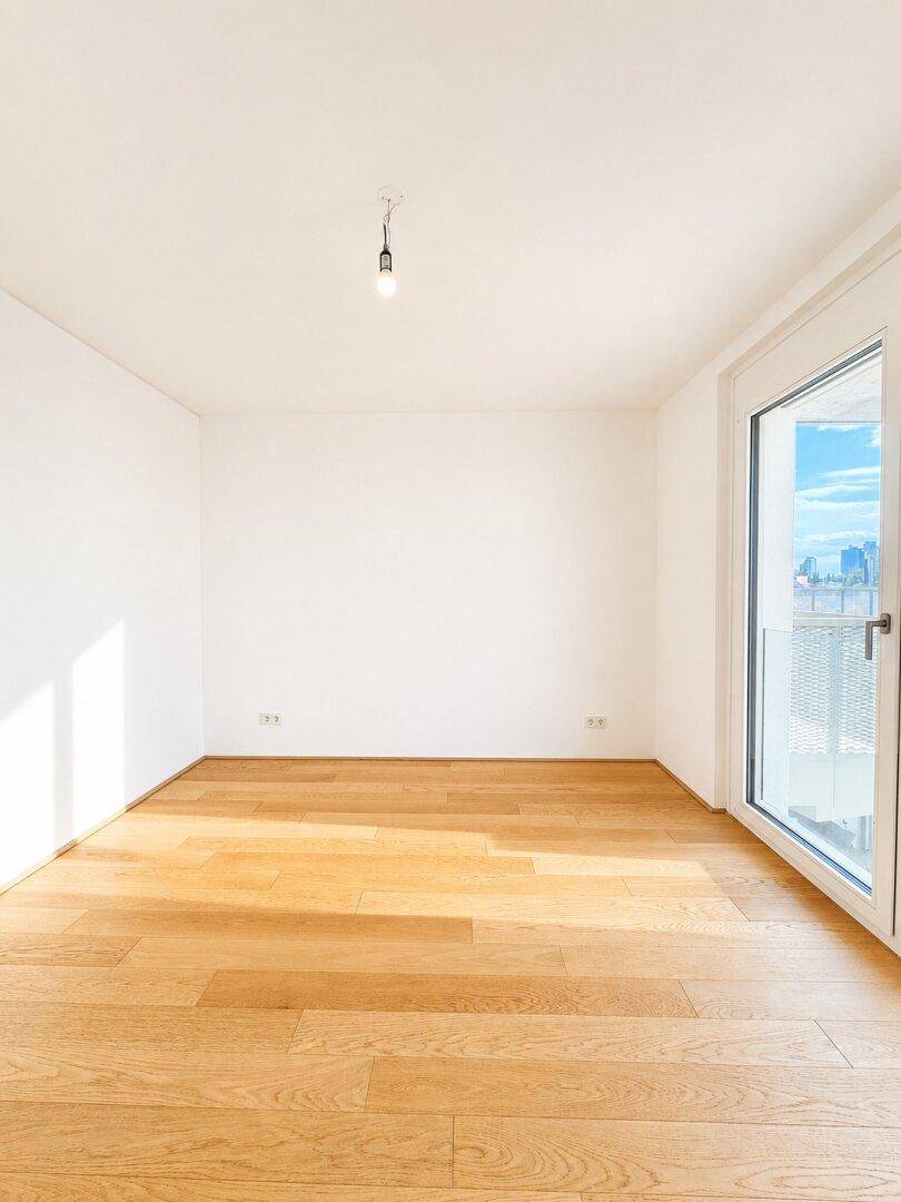 Helles, leeres Zimmer mit Holzboden und einer Fenstertür, die viel Tageslicht hereinlässt.