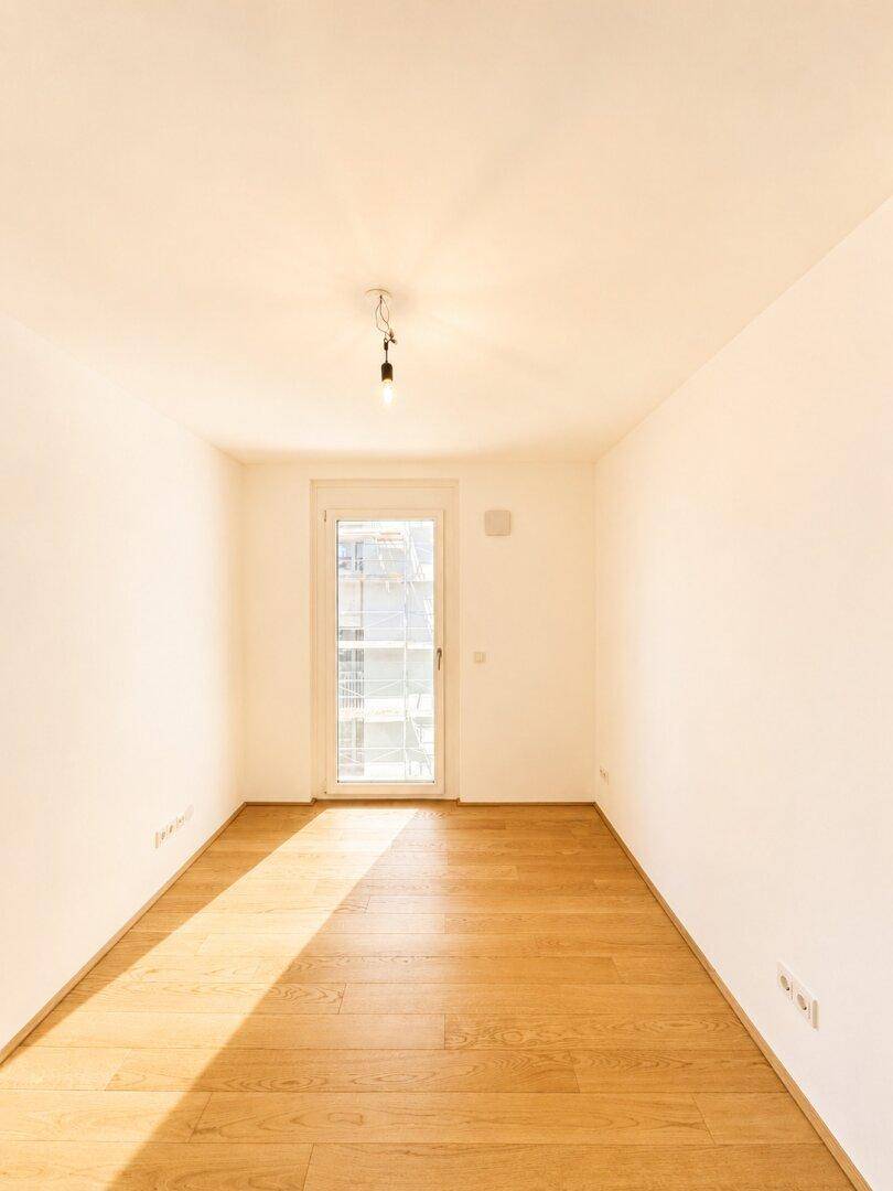 Leeres, sonniges Zimmer mit Holzboden und einer Fenstertür, die den Raum gut belichtet.