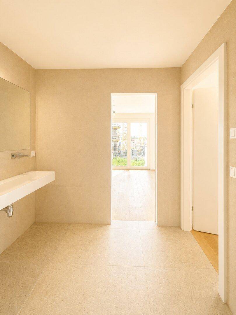 Modernes Badezimmer mit beigefarbenen Fliesen und Blick in einen hellen Raum mit Zugang nach draußen.