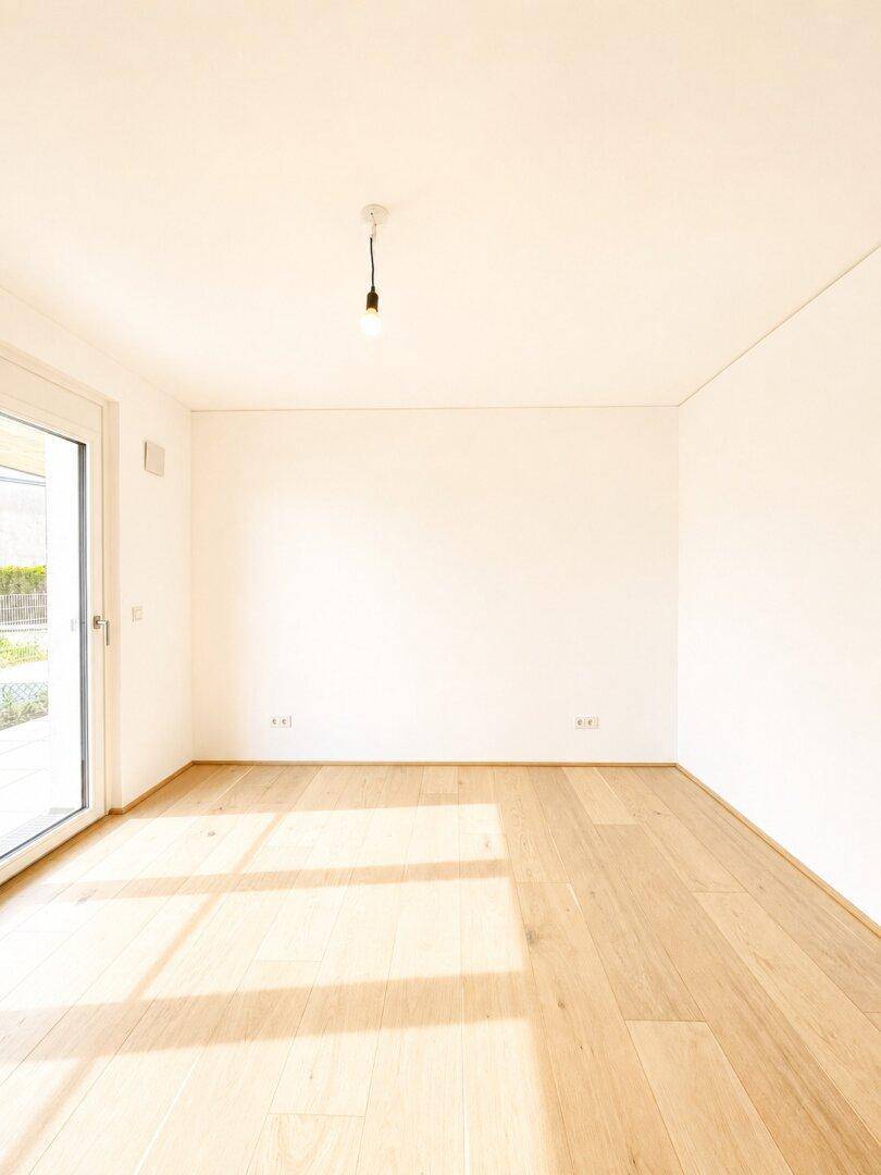 Helles, leeres Zimmer mit Holzboden und Fenster, das viel Tageslicht hereinlässt.