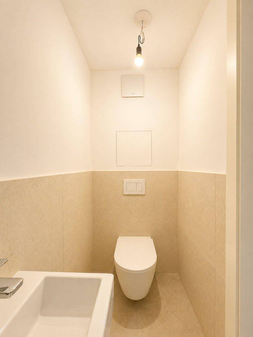 Modernes Gäste-WC mit Wand-WC, kleinem Waschbecken und beigefarbenen Fliesen.