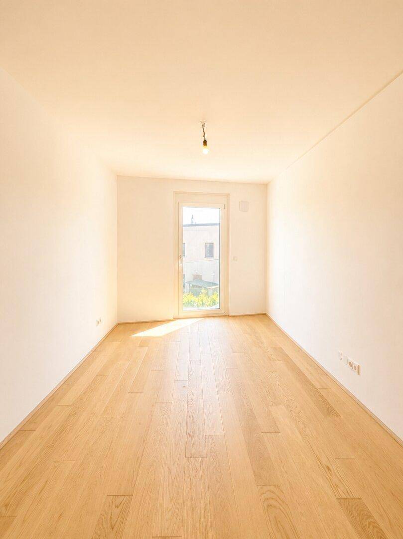 Leeres Zimmer mit Holzboden und einem großen Fenster, das den Raum erhellt.