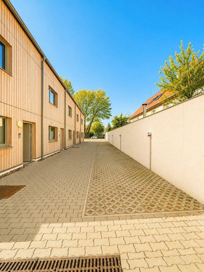 Gepflasterter Weg zwischen Reihenhäusern und einer weißen Mauer, mit viel Sonnenlicht.