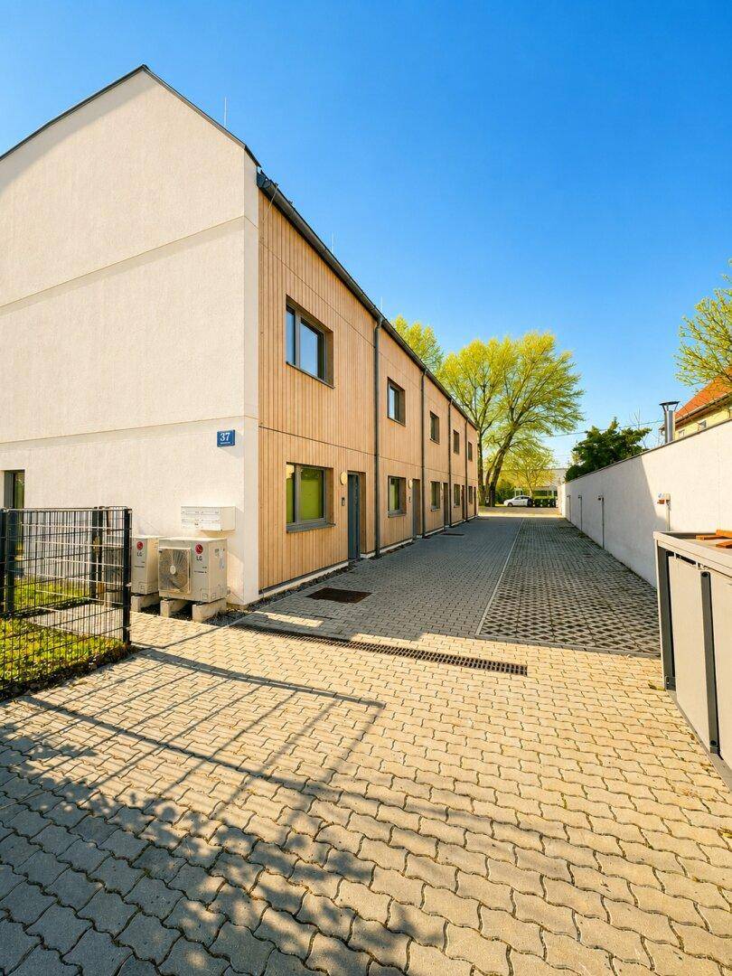 Moderne Reihenhäuser mit Holz- und Putzfassade sowie gepflastertem Zugangsbereich und Wärmepumpen.