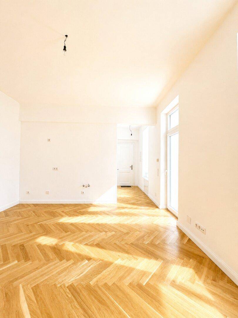 Lichtdurchfluteter Raum mit klassischem Fischgrätparkett und Zugang zu einem Flur, bereit zum Einrichten.