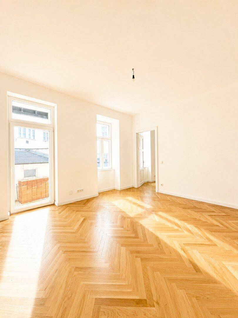 Großes, helles Zimmer mit Fischgrätparkett und Zugang zu einem Balkon, das viel Tageslicht bietet.
