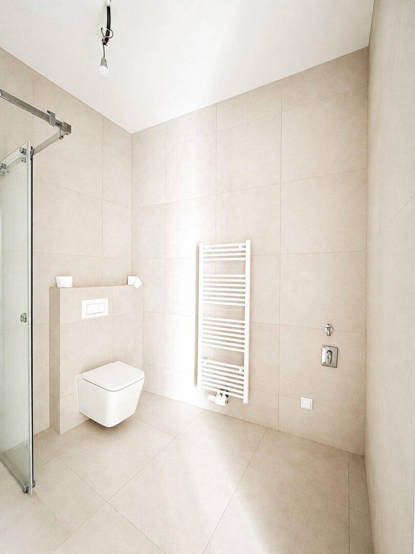 Modernes Badezimmer mit beigefarbenen Fliesen, wandhängendem WC, Dusche und beheiztem Handtuchhalter.