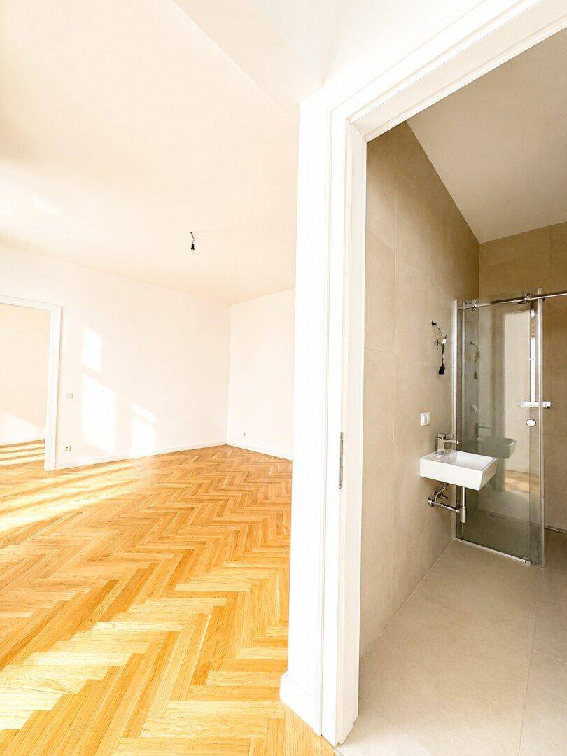 Blick von einem hellen Zimmer mit Fischgrätparkett in ein modernes Badezimmer mit Dusche.