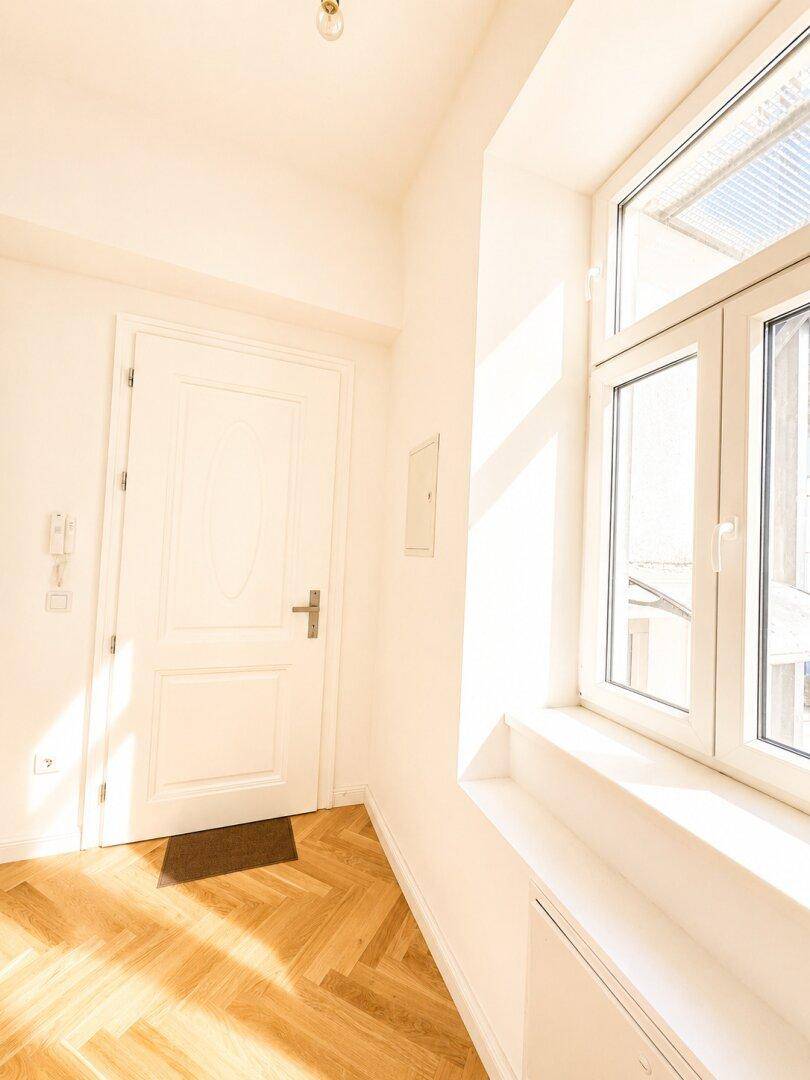 Heller Eingangsbereich mit Fischgrätparkett, weißer Tür und Fenster, das Tageslicht hereinlässt.