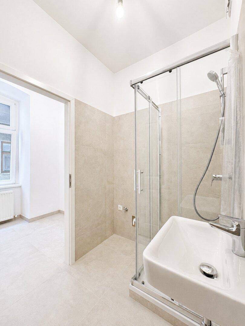 Modernes Badezimmer mit Duschkabine und Waschbecken, mit Blick in einen hellen Nebenraum.