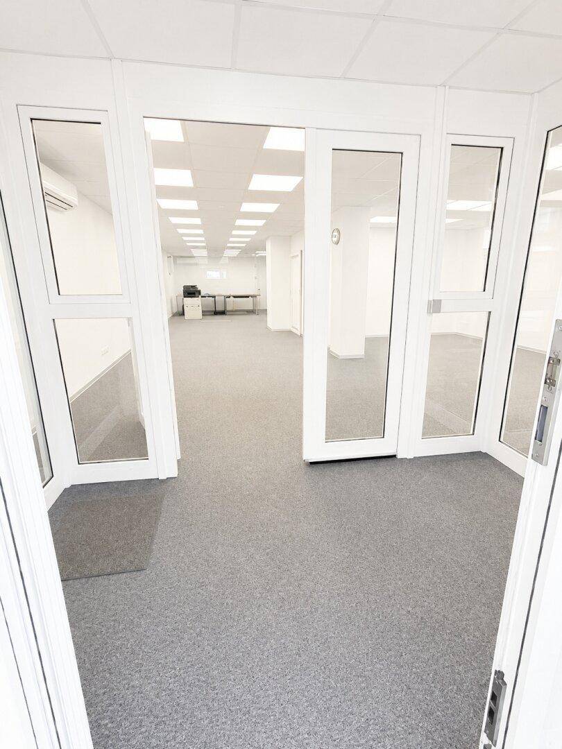 Eingangsbereich eines Büros mit Glastüren, grauem Teppichboden und Blick in den Hauptraum.