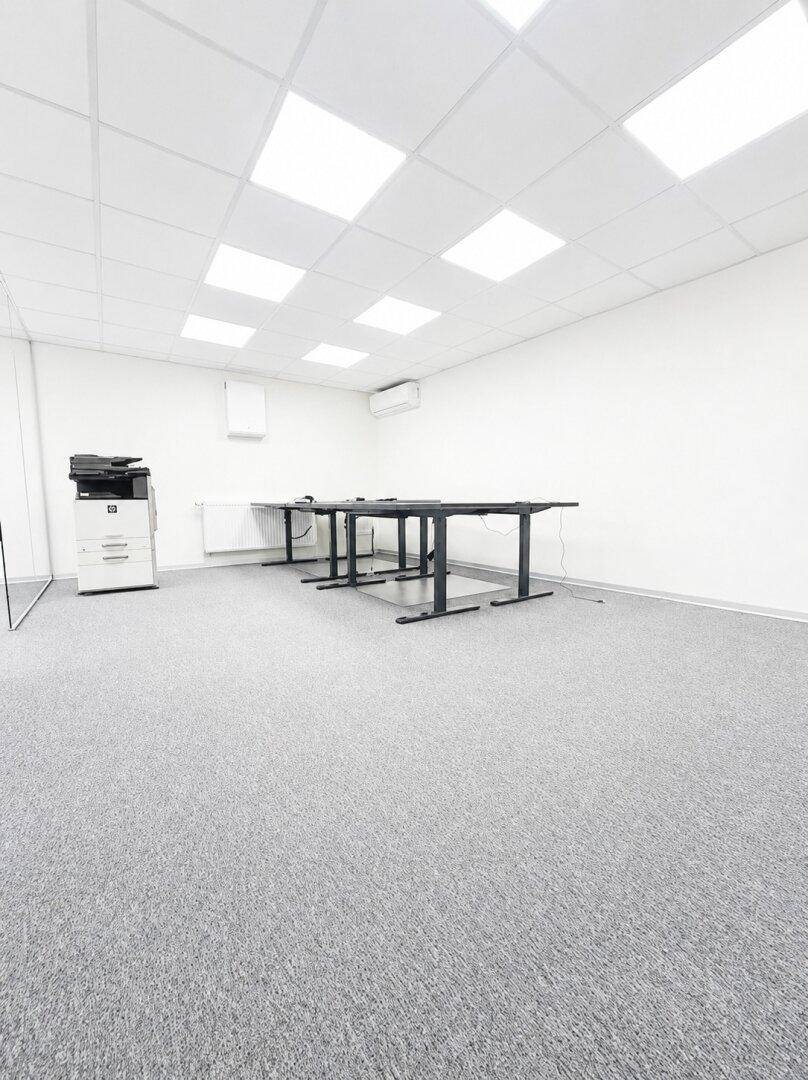 Bürofläche mit grauem Teppichboden, weißen Wänden und einem Kopierer, bereit zur Nutzung.