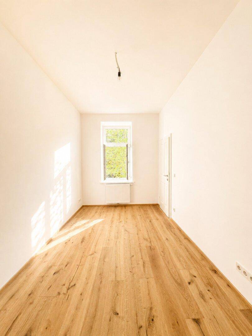 Langes, helles Zimmer mit einem Fenster und neu verlegtem Holzboden, ideal als Schlafzimmer.