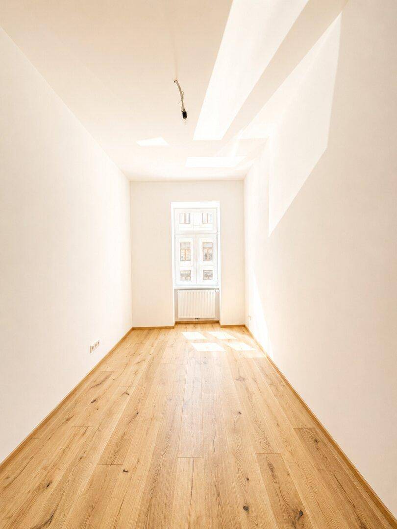 Schmaler, heller Raum mit Holzboden und einem Fenster, das Tageslicht spendet.