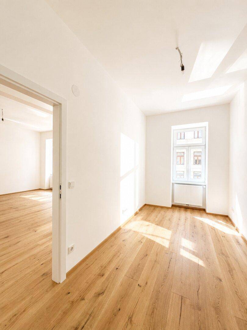 Heller Wohnbereich mit Holzboden und Zugang zu einem weiteren Zimmer, durchflutet von Sonnenlicht.