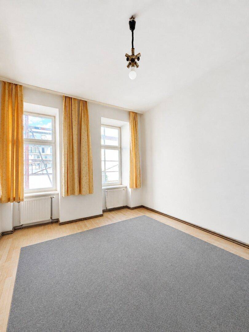 Helles, leeres Zimmer mit Holzboden, zwei Fenstern mit gelben Vorhängen und einfacher Deckenleuchte.