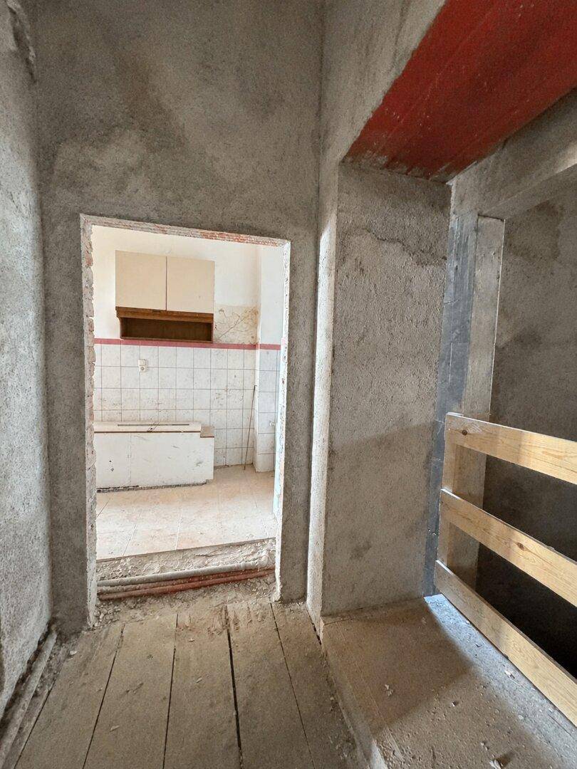 Flur im Rohbauzustand mit unverputzten Wänden und sichtbaren Holzbalken, führt zu einem gefliesten Raum.