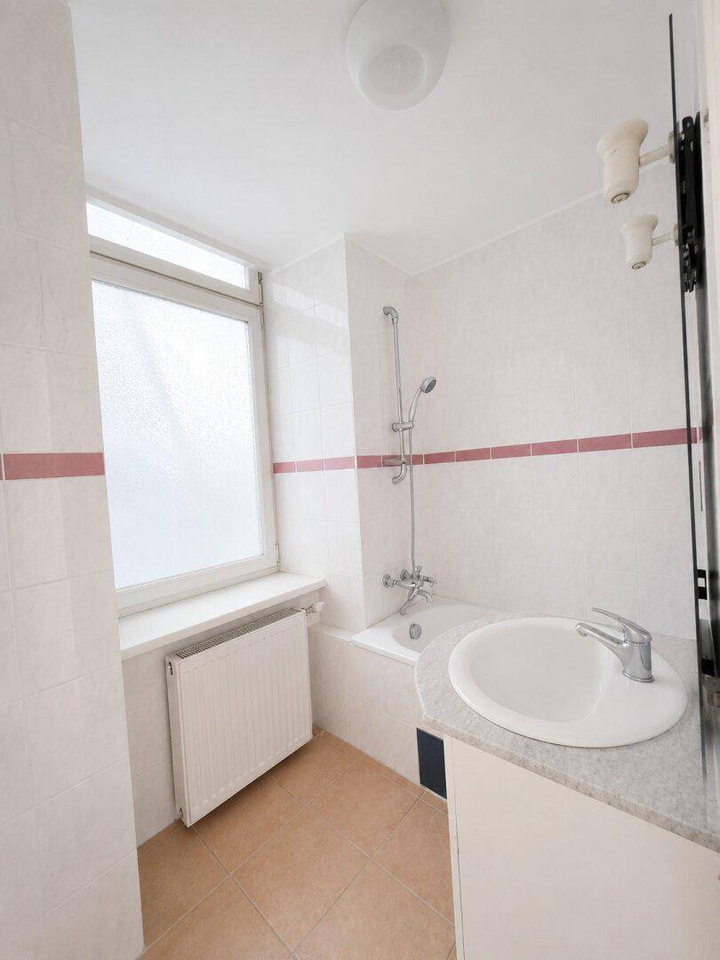 Helles Badezimmer mit Badewanne, Dusche, Waschbecken und Fenster, ausgestattet mit einem Heizkörper.