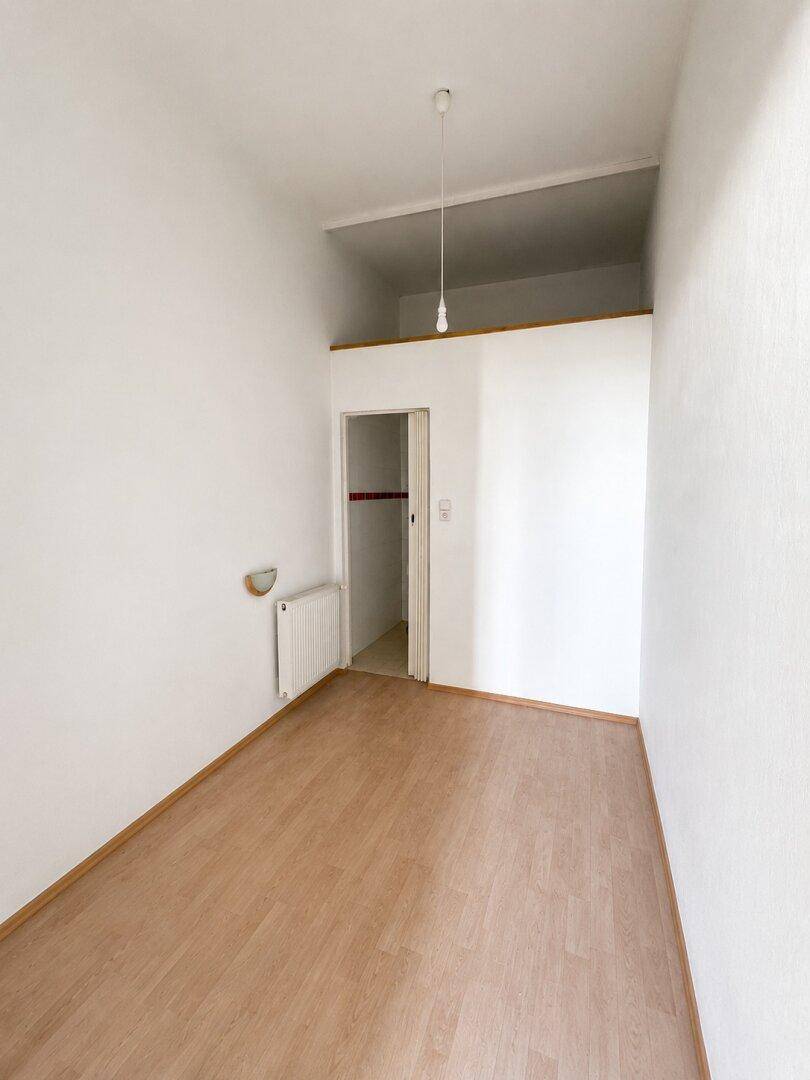 Leeres Zimmer mit hellem Holzboden, weißen Wänden und Heizkörper, inklusive kleinem begehbarem Schrankbereich.