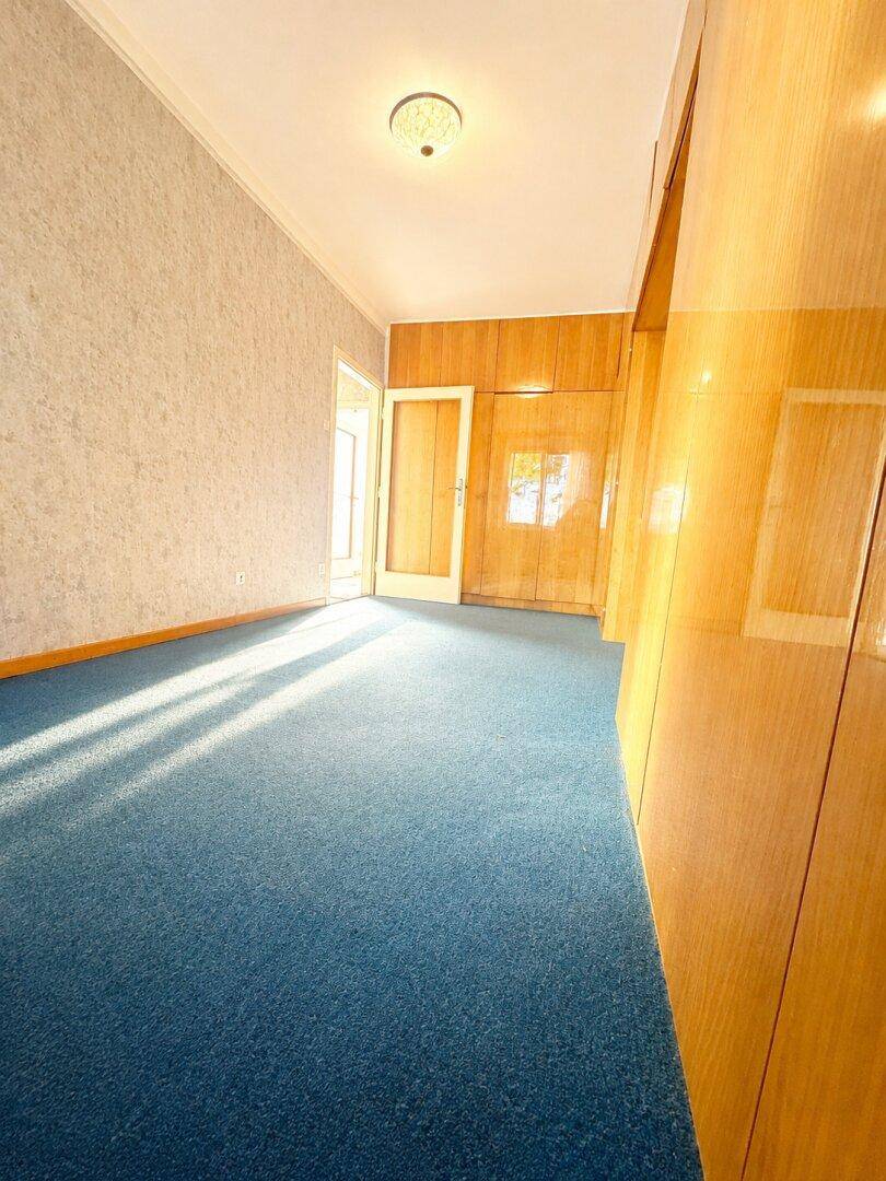 Schlafzimmer mit blauem Teppichboden und eingebauten Holzschränken, hell beleuchtet durch die Sonne.