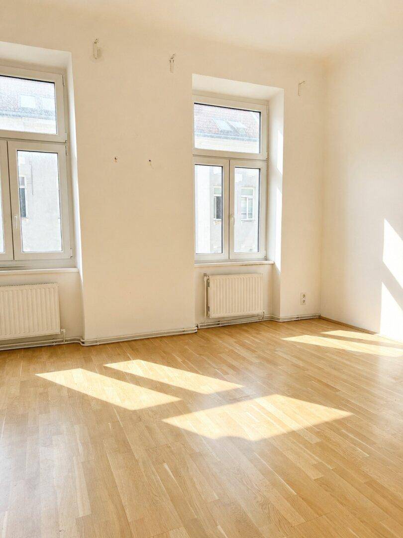 Lichtdurchflutetes Zimmer mit Holzboden und zwei Fenstern, die viel Tageslicht hereinlassen.