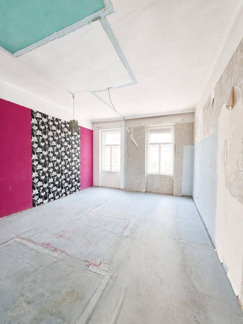 Renovierungsbedürftiges Zimmer mit freigelegten Wänden, Betonboden und farbigen Akzenten an Decke und Wand.