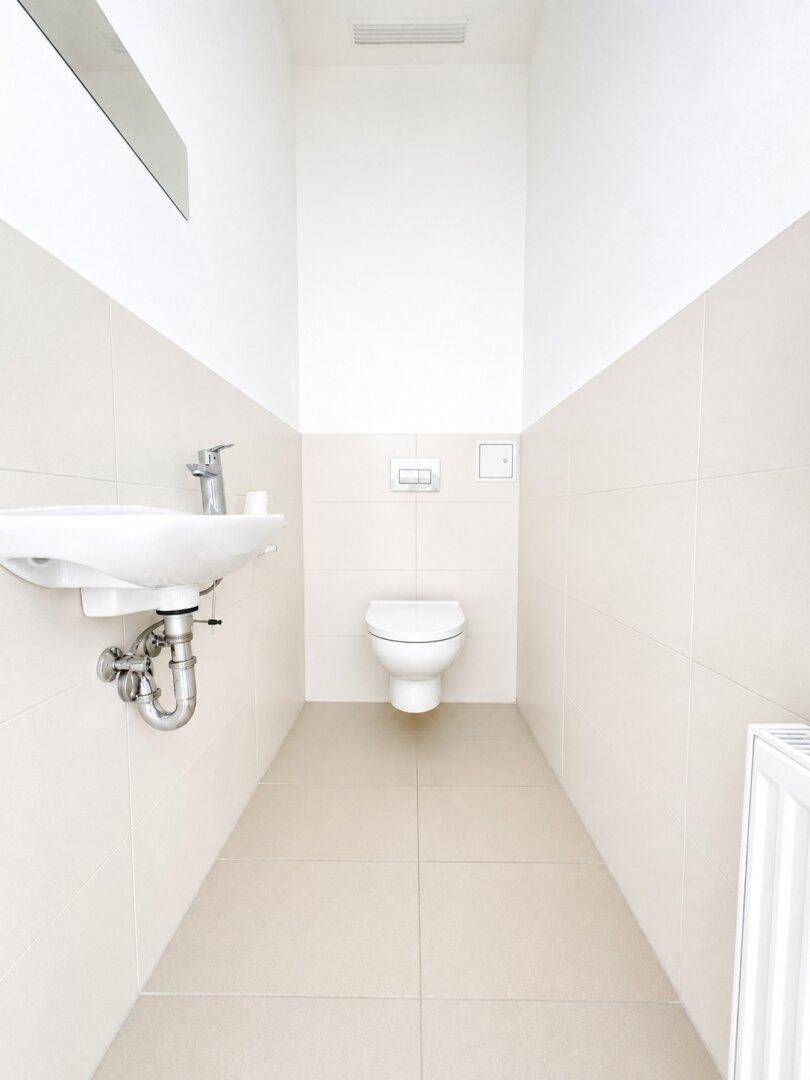 Separates WC mit wandhängender Toilette, Waschbecken und hellen Fliesen.